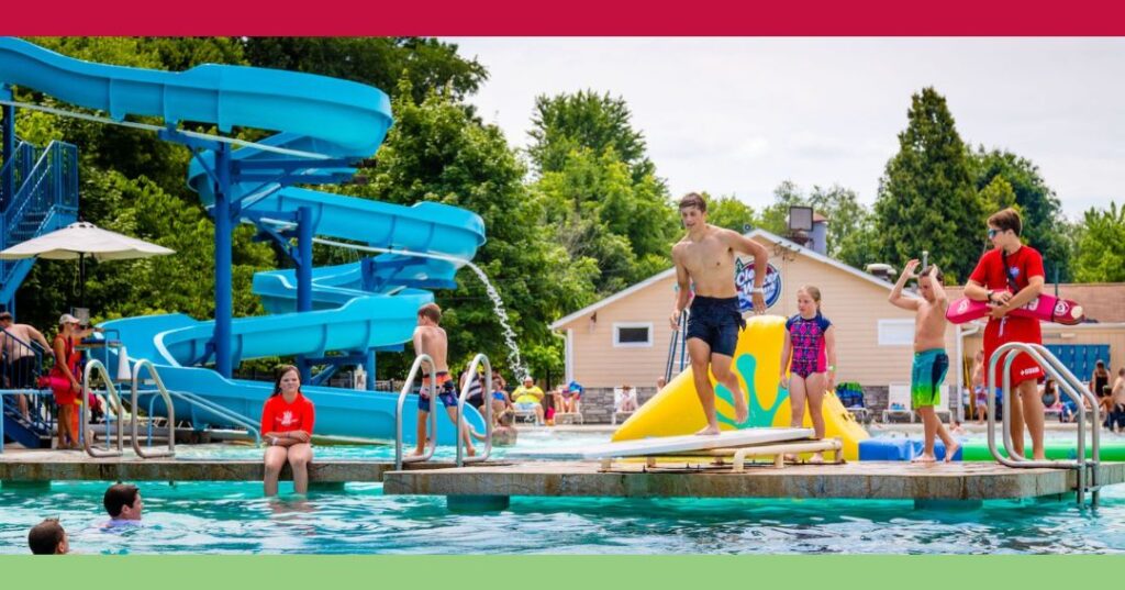 Children and teenagers play on a water slide and floating obstacle course in an outdoor swimming pool on a sunny day.