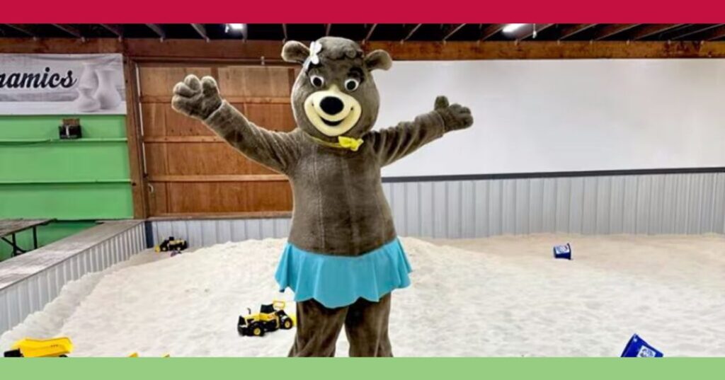 A person in a bear costume with a blue skirt stands with arms outstretched in a large indoor sandbox area with toy trucks.