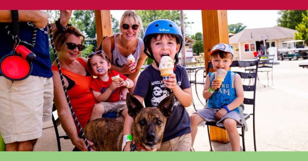 A group of people and a dog sit outdoors eating ice cream; a child wearing a helmet is at the front, smiling at the camera.