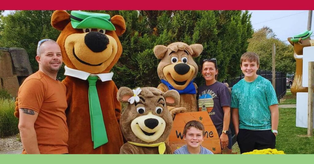 A family poses outdoors with three people in bear costumes, including Yogi Bear and Boo Boo, in front of greenery and a “Welcome” sign.