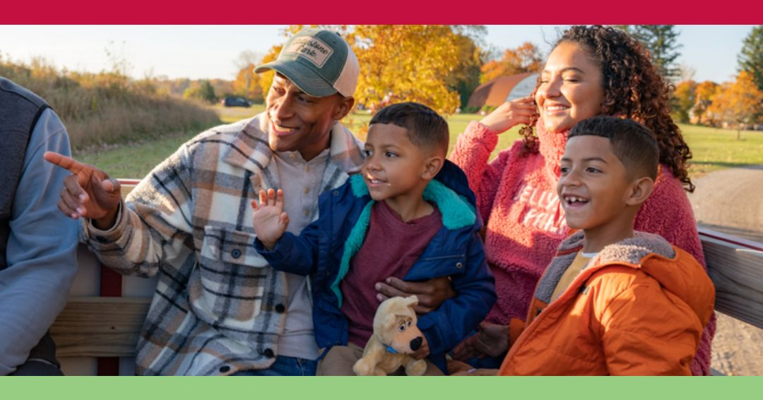 A family of four sits on a wooden bench outdoors, smiling and waving on a sunny autumn day; colorful trees and a barn are visible in the background.