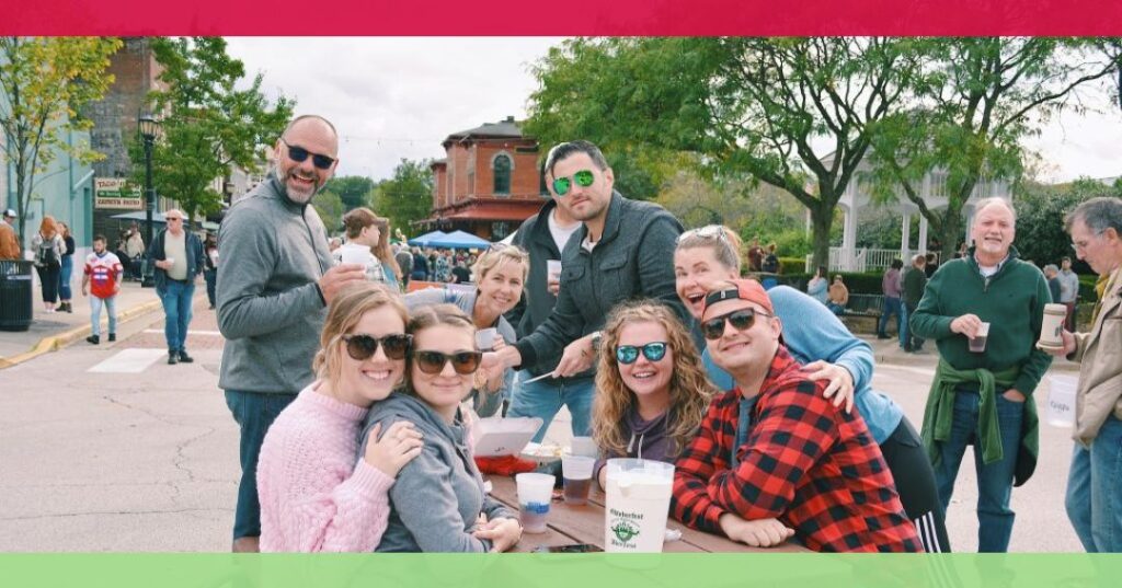 A group of people wearing sunglasses gather around a table outdoors at a street event, smiling at the camera with food and drinks on the table.