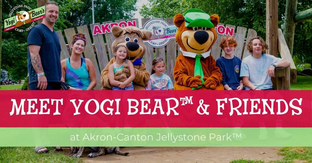 A family poses with people in Yogi Bear™ and Boo Boo™ costumes at Akron-Canton Jellystone Park™, with a wooden bench and park signs in the background.