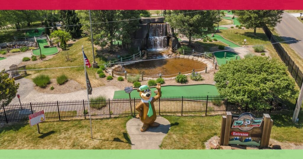 Aerial view of a mini golf course with a large Yogi Bear statue at the entrance, a pond with a waterfall, and surrounding greenery.