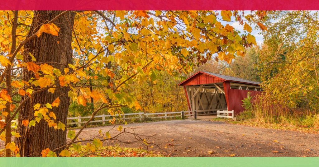 A red covered bridge surrounded by autumn trees with yellow leaves, a dirt path, and a white wooden fence.