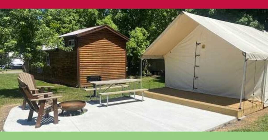 A campsite with a wooden cabin, a canvas tent on a platform, a picnic table, Adirondack chairs, and a fire pit on a concrete pad, surrounded by trees.
