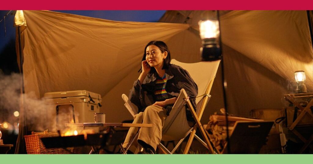 A person sits in a camping chair under a tent at night, talking on the phone, with lanterns and a fire pit nearby.