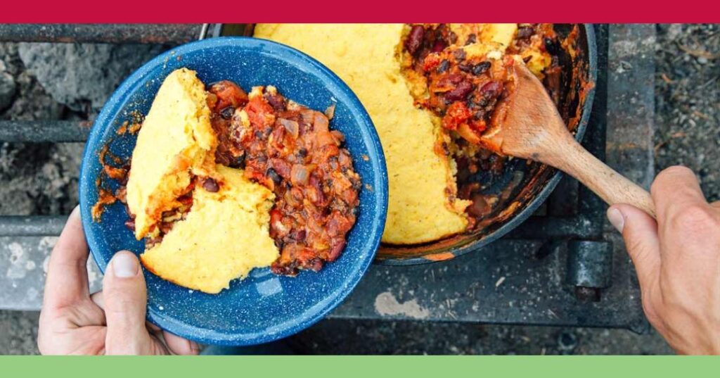 A person serves a portion of chili with cornbread topping from a cast iron skillet onto a blue plate using a wooden spoon.