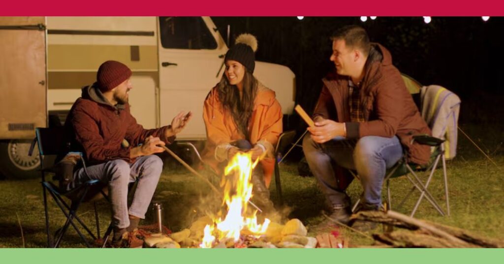 Three people in jackets sit around a campfire at night, talking and smiling, with a camper van and string lights in the background.