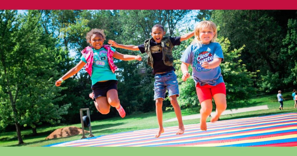 Three children are jumping and smiling on an inflatable mat outdoors, with green trees and grass in the background on a sunny day.