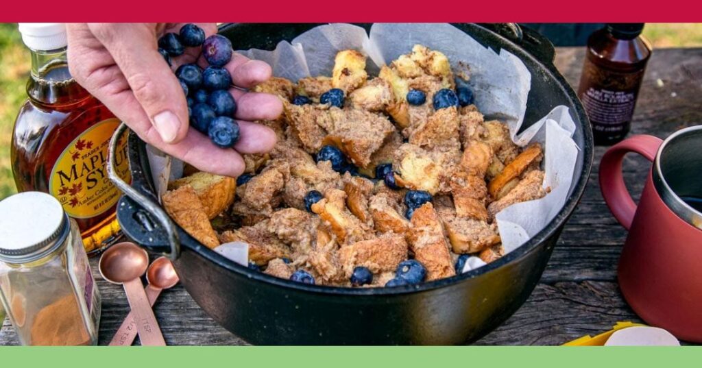A hand sprinkles fresh blueberries over a bread and cinnamon mixture in a black pot, surrounded by maple syrup, cinnamon, measuring spoons, and a mug on a wooden table.