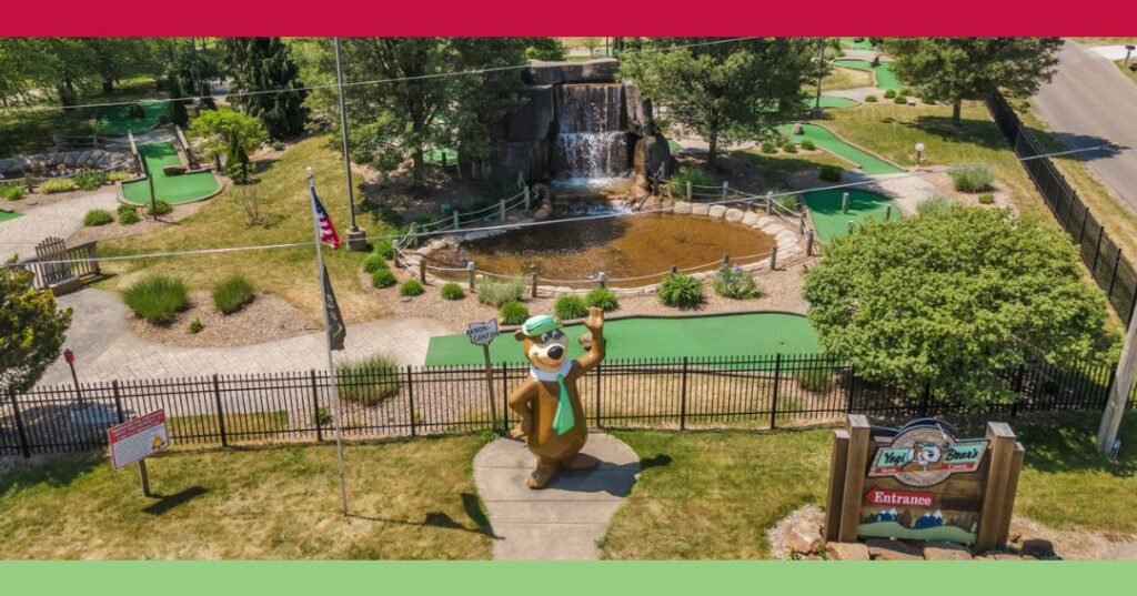 A mini-golf course with a Yogi Bear statue at the entrance, surrounded by greenery, a pond with a waterfall, and an American flag.