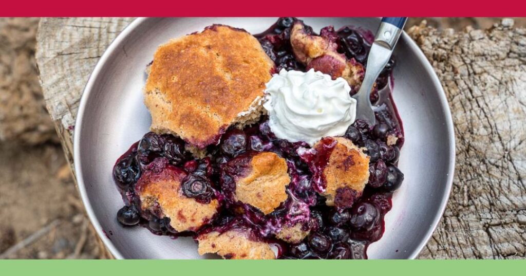 A plate of blueberry cobbler topped with a dollop of whipped cream, served with a spoon on a wooden surface.