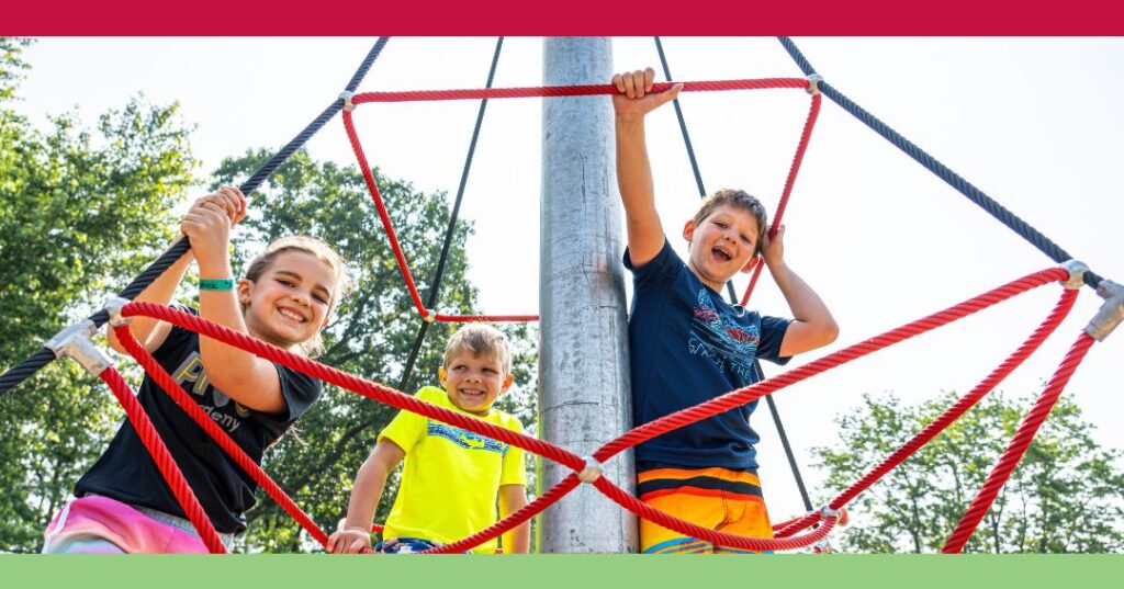 Three children climb and smile on a red rope play structure outdoors, with trees and a metal pole in the background.