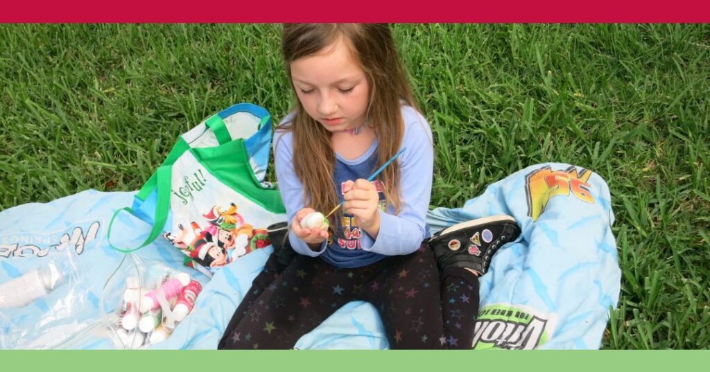 A young girl sits on a blanket in the grass, examining an object in her hands, with art supplies and a tote bag beside her.
