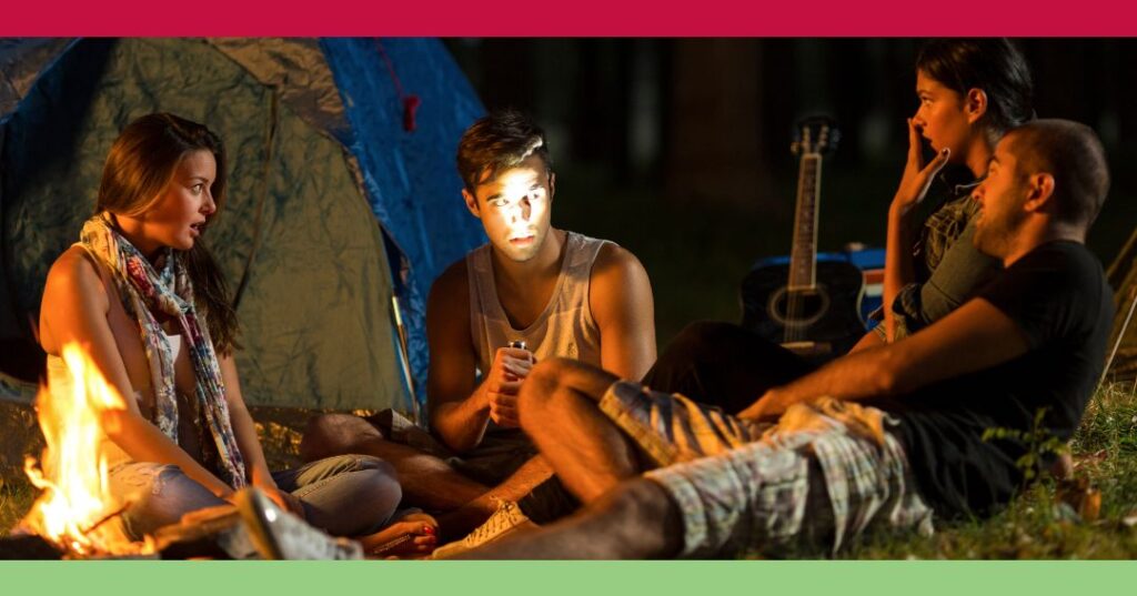 Four people sit in a circle at a campsite at night, one holding a flashlight under their face, with a tent and a guitar in the background.