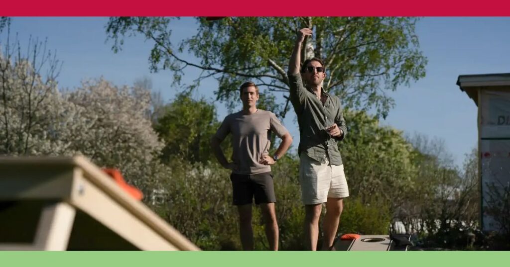 Two men play cornhole outdoors; one is tossing a beanbag while the other stands with hands on hips. Trees and a building are visible in the background.