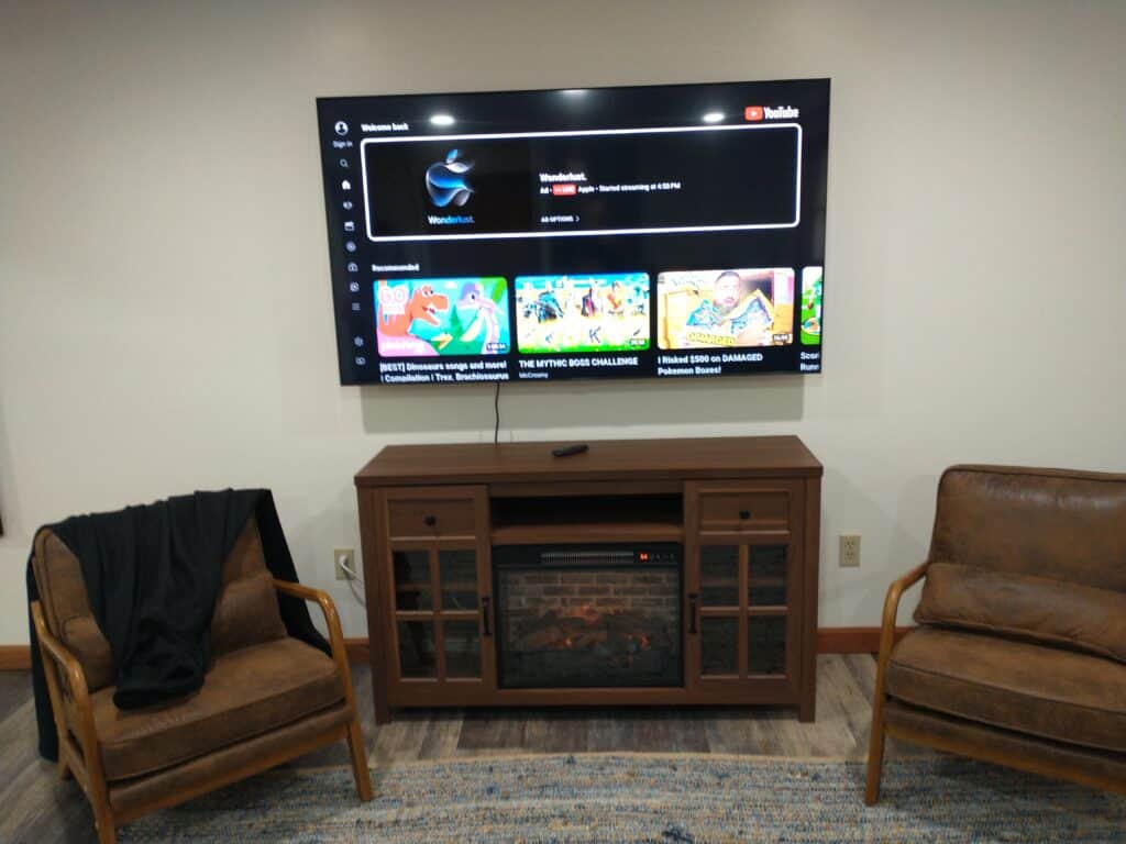 Wall-mounted TV displaying YouTube above a wooden electric fireplace, flanked by two brown chairs, one with a black jacket draped over the arm.