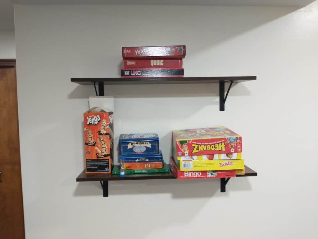 Two wall-mounted shelves hold various board games, including Jenga, Scrabble, Pictionary, UNO, Qubic, Taboo, Headbanz, and Bingo, against a plain white wall.