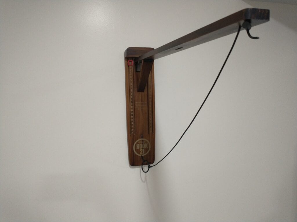 A wooden wall-mounted hook game with a string and ring attached, and a horizontal arm extending outward, against a plain white wall.