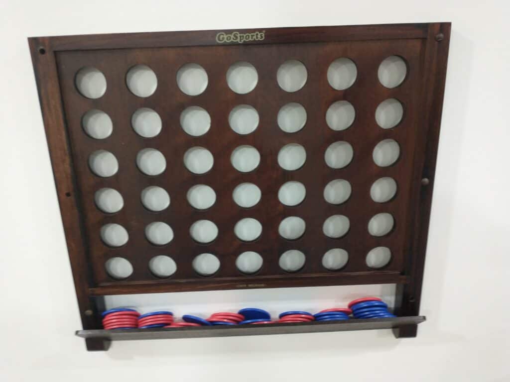 A wooden Connect Four game board is mounted on a wall, with red and blue discs collected in a tray at the bottom.