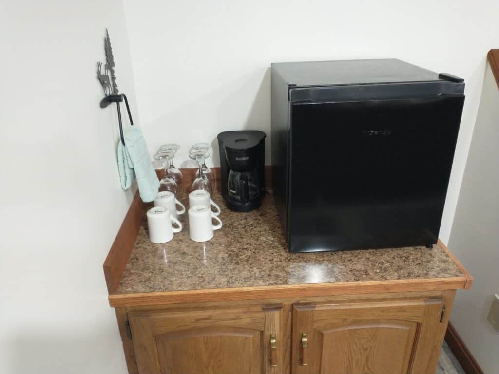 A countertop with a mini fridge, coffee maker, three white mugs, four upside-down glasses, and an oven mitt hanging on the wall above.