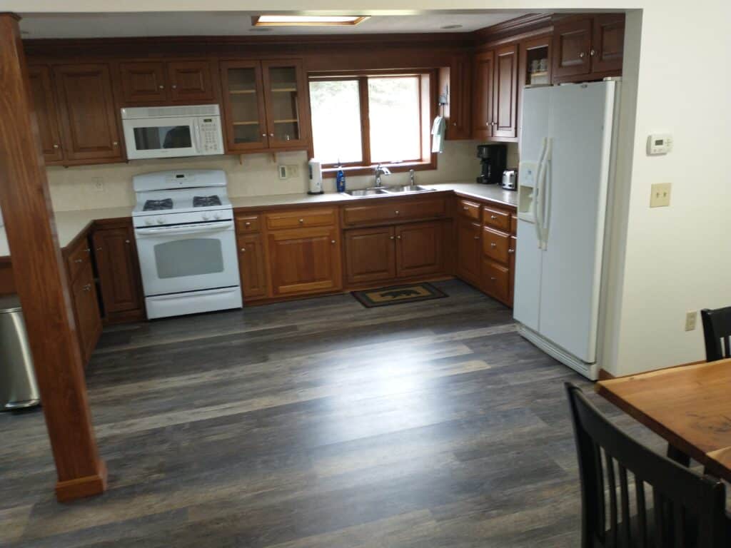 Modern kitchen with wooden cabinets, white appliances, a double-door refrigerator, a microwave, and a window above the sink. Dark wood dining table and chairs are partially visible.