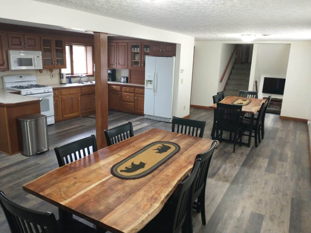 Spacious kitchen and dining area in cozy cabins, featuring wood cabinets, two wooden tables with bear designs, black chairs, and a view of stairs and the living room in the background.