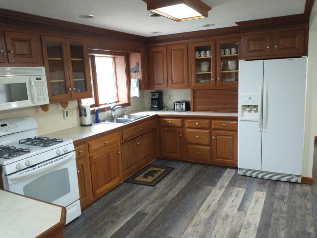 A kitchen with wooden cabinets, white appliances including a gas stove, microwave, and refrigerator, and a window above the sink. The floor is wood-patterned vinyl.