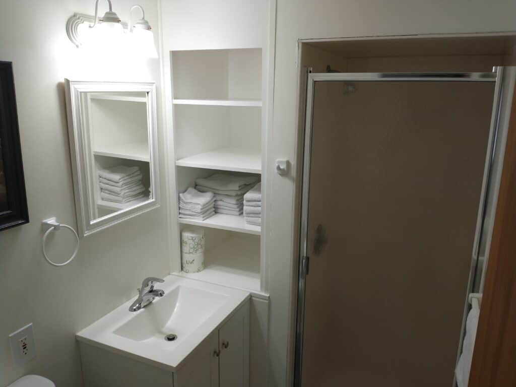 Small bathroom with a white sink, mirror, open shelves holding folded towels, a wall-mounted light, and a glass shower enclosure.