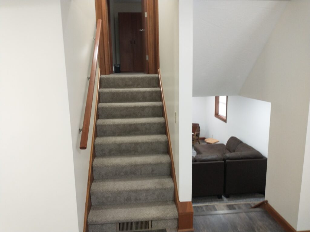 Carpeted staircase with a wooden handrail leading up to a closed door; to the right, a living area with brown sofas and a small table is partially visible.