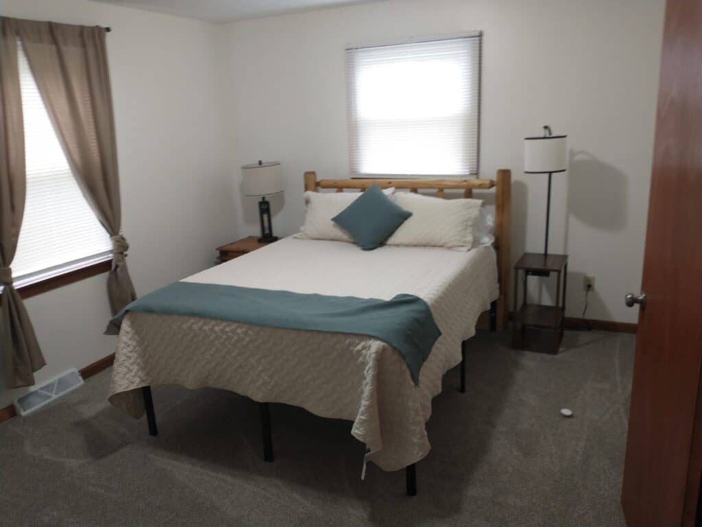 A small bedroom with a double bed, two bedside tables with lamps, beige walls, carpeted floor, and two windows with curtains.