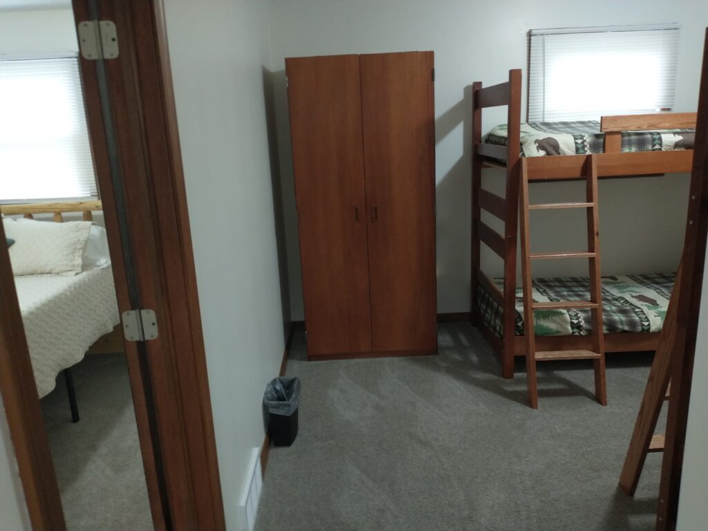 A small bedroom with a wooden wardrobe, a bunk bed, and a window; another room with a bed is visible through an open doorway.