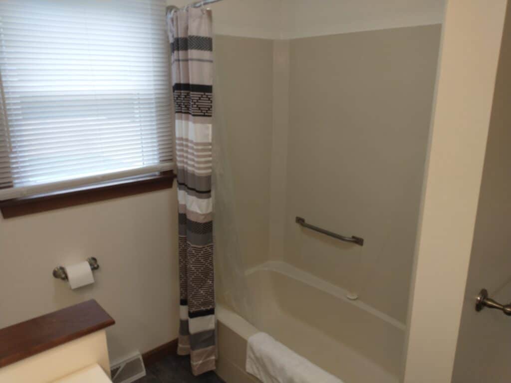 Bathroom with a bathtub and shower combination, striped shower curtain, towel on the tub edge, grab bar, toilet paper holder, and window with blinds.