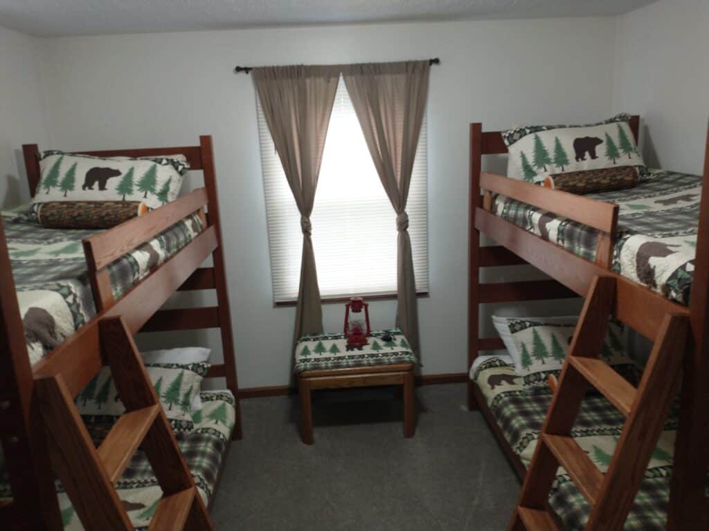 A small bedroom with two wooden bunk beds, nature-themed bedding, a window with beige curtains, and a bench beneath the window.
