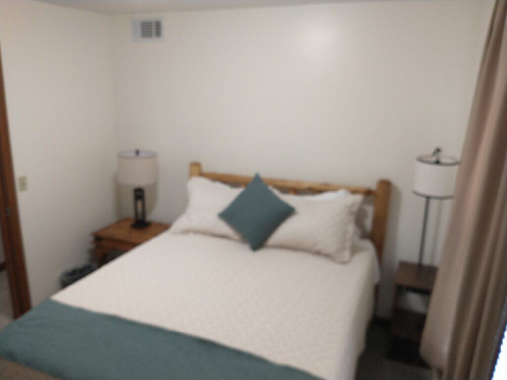 A small bedroom with a bed centered against a white wall, two lamps on nightstands, and light-colored bedding with blue accents.