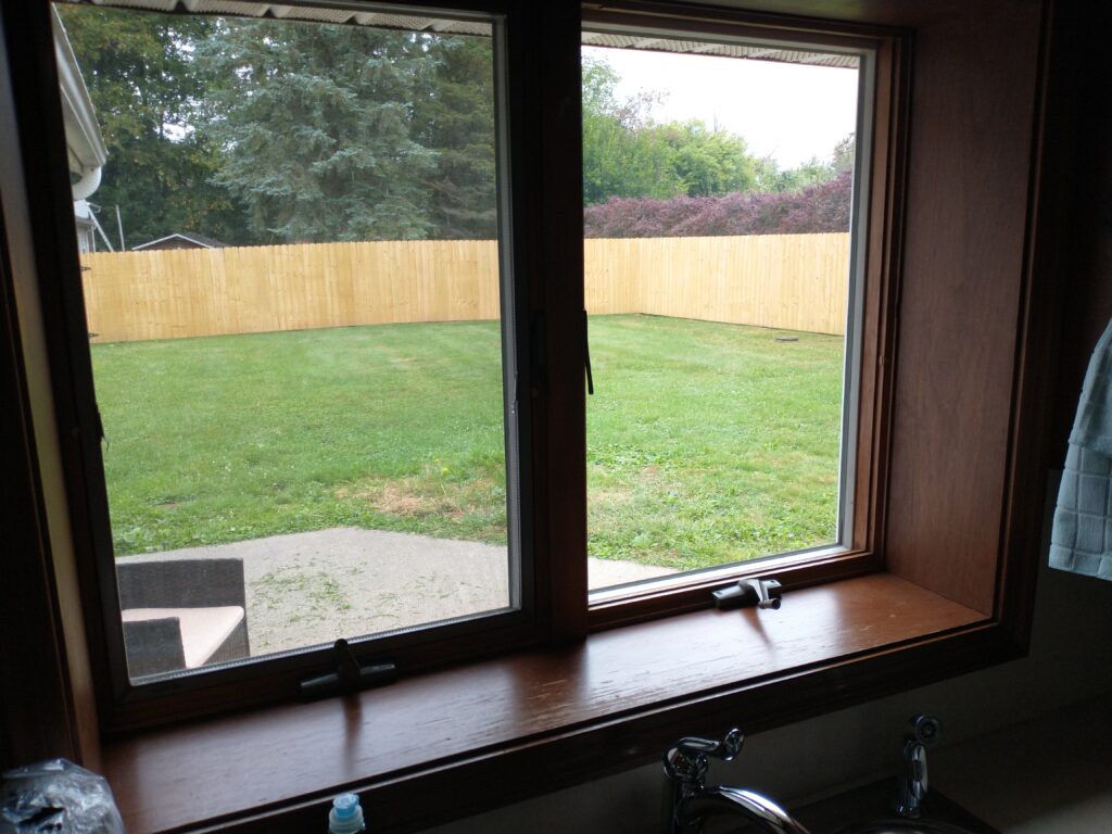 A view through a window shows a green backyard enclosed by a wooden fence, with a patio and part of a sink visible inside.