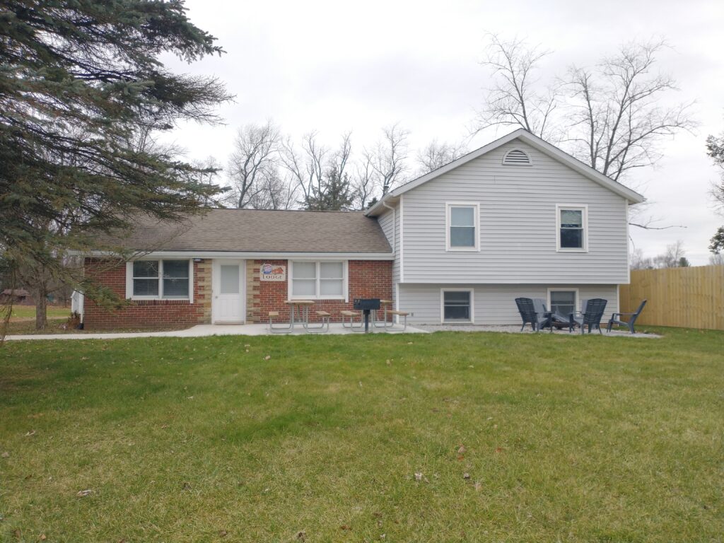 A split-level house with brick and light-colored siding, a small patio with tables and chairs, and a grassy backyard. Trees without leaves are visible in the background.