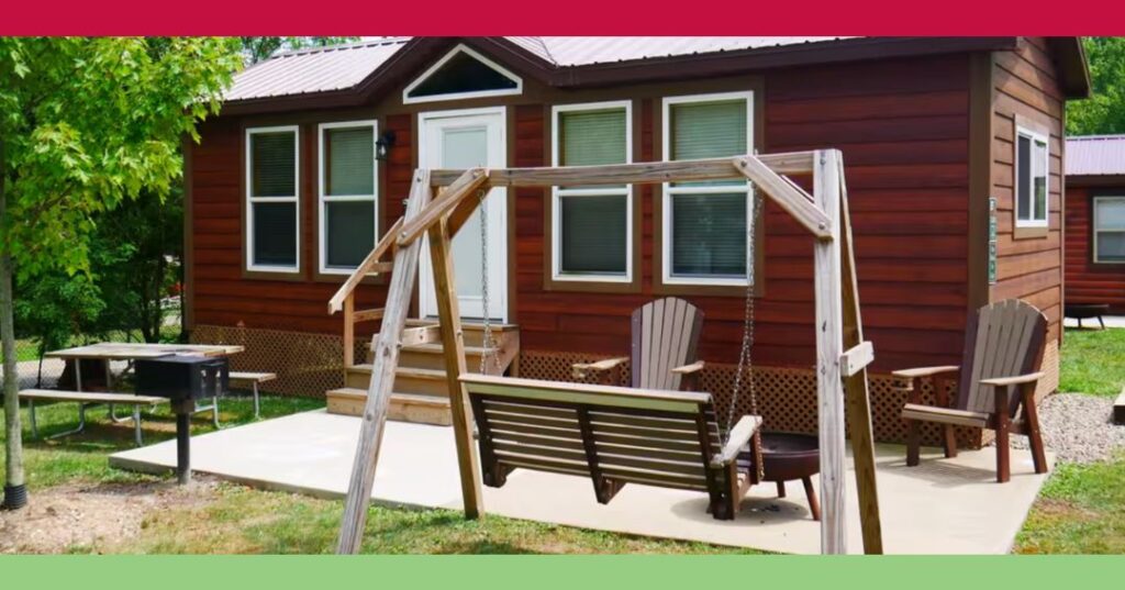 A small wooden cabin with a front porch, a wooden swing, two chairs, a picnic table, and a grill on a concrete patio in a green outdoor setting.