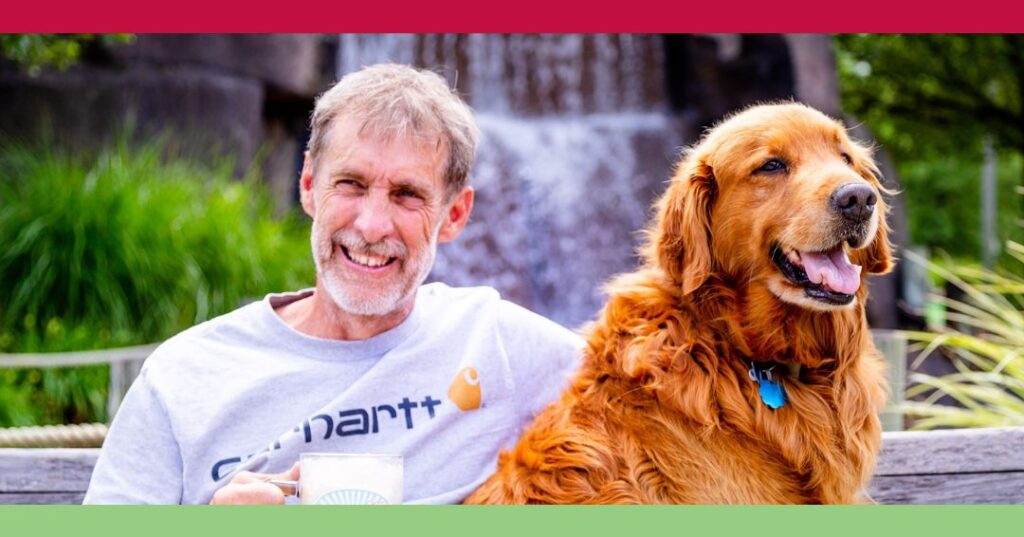 A man with gray hair and a beard sits on a bench outdoors, smiling and holding a mug, next to a happy golden retriever. A waterfall is visible in the background.