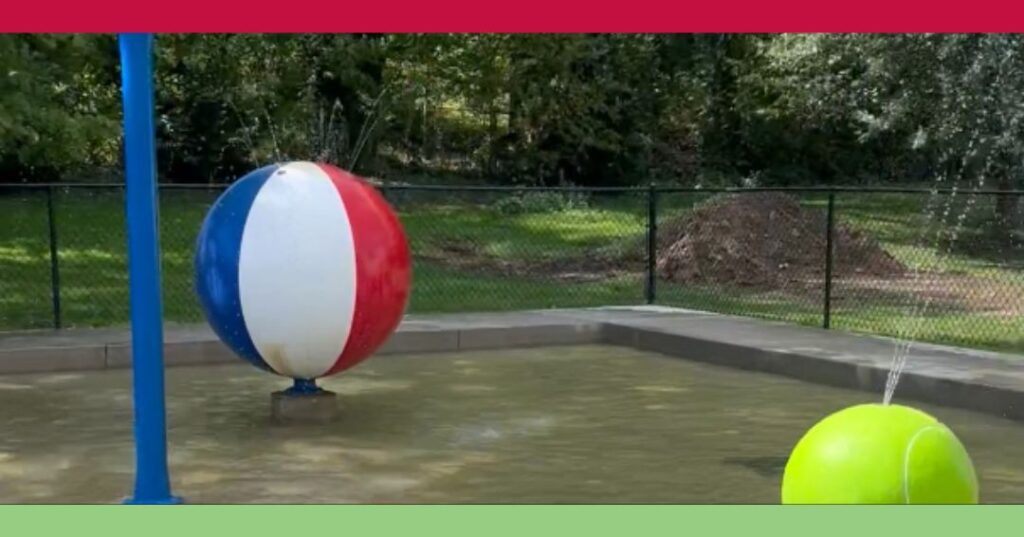 A splash pad features a large beach ball and a tennis ball spraying water, surrounded by a fence and greenery in the background.