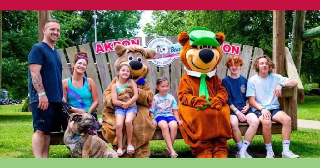 A group of people, two children in bear costumes, and a dog sit on a large wooden bench at Akron, Ohio's Jellystone Park, with trees and grass in the background.
