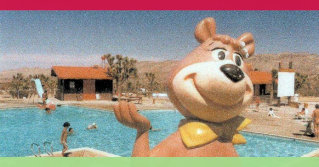 A large Yogi Bear statue stands by a swimming pool with children swimming; buildings and desert landscape are visible in the background.