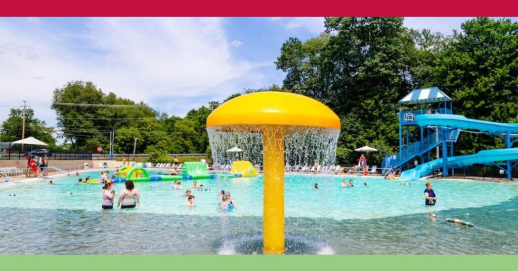 Outdoor public pool with people swimming, a yellow mushroom-shaped water feature in the center, and a blue water slide on the right, surrounded by trees and umbrellas.
