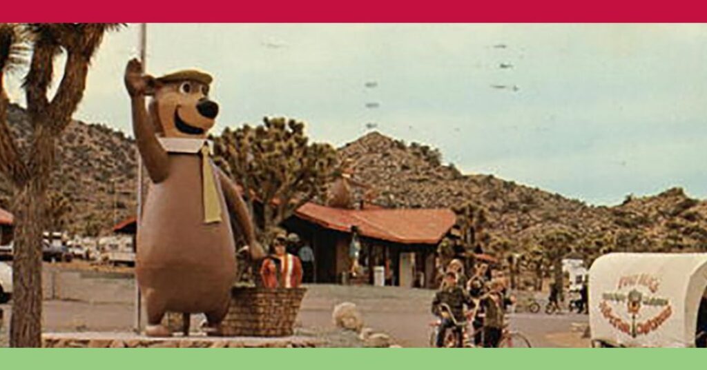 A large statue of Yogi Bear stands beside a flagpole in front of a building, with children on bicycles and a covered wagon nearby in a desert landscape.