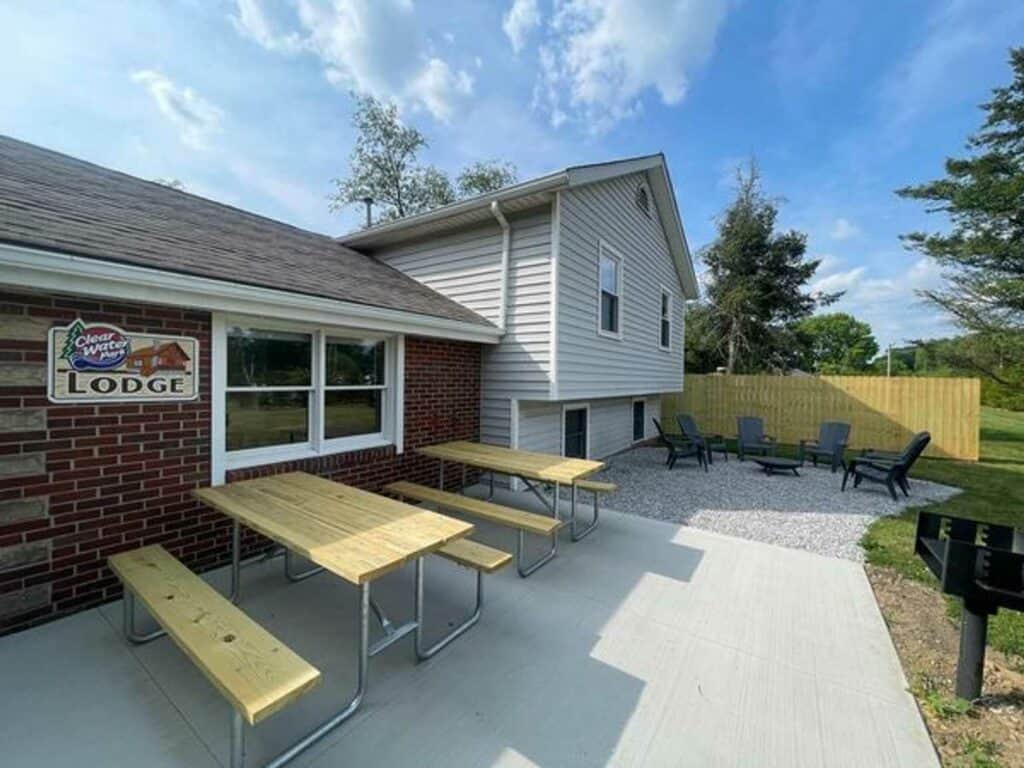 Outdoor area with picnic tables, a grill, and a seating area with chairs on gravel beside a brick and siding building labeled “Lodge.” Blue sky and green trees in the background.