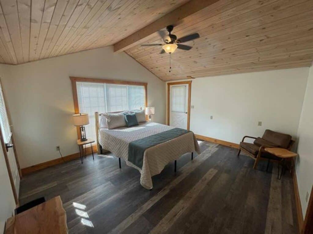 A simple bedroom with a wooden ceiling, queen bed, two bedside lamps, an armchair with ottoman, large windows, and dark wood flooring.