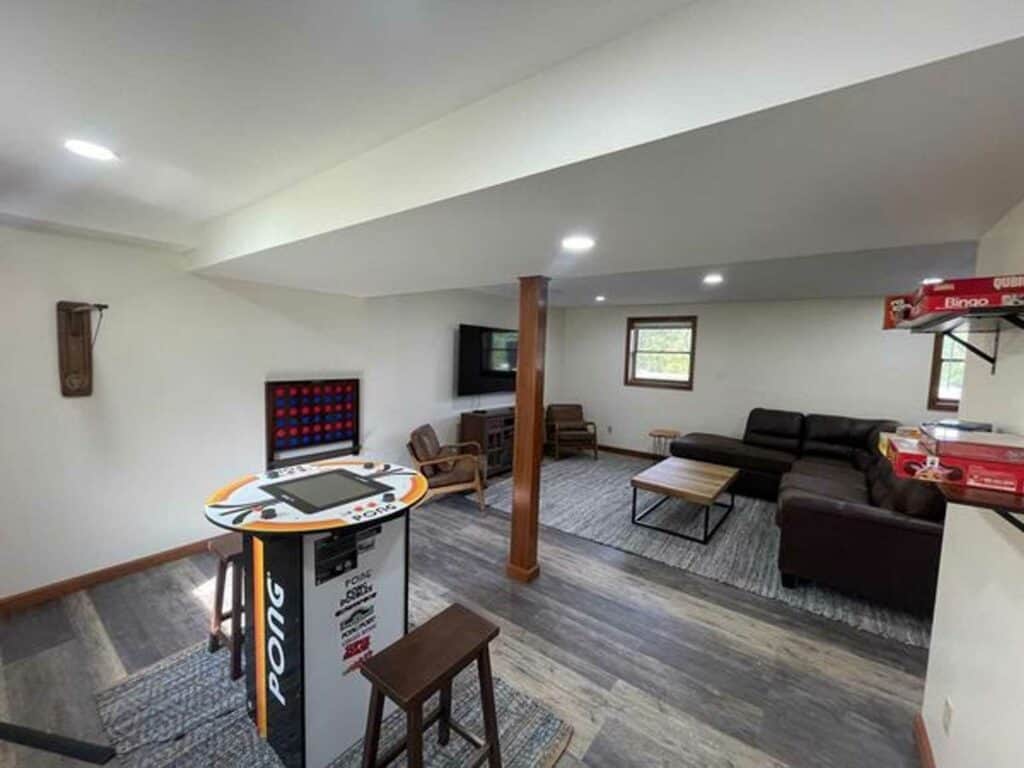 A basement game room in cozy cabins featuring an arcade machine and stool, Connect 4, board games on shelves, a sectional sofa, armchairs, a TV, and windows.