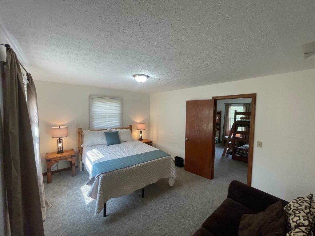 A cozy bedroom in one of the cabins features a bed, two side tables with lamps, a window, an open door to a room with bunk beds, and a couch in the foreground.