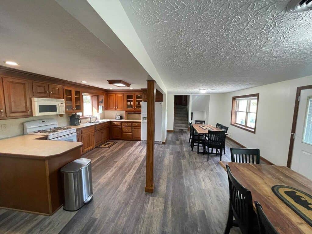 Spacious open-plan kitchen and dining area with wooden cabinets, modern appliances, two tables, and large windows letting in natural light.